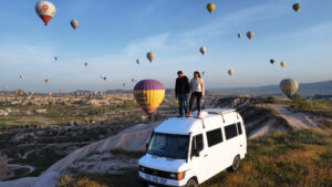 A couple stands on a van, smiling as colorful hot air balloons float in the sky above them.