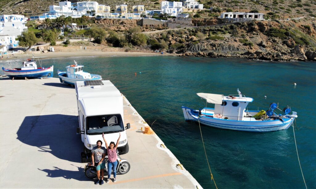 A couple standing on a dock near a white campervan, with a beach and houses visible in the background.
