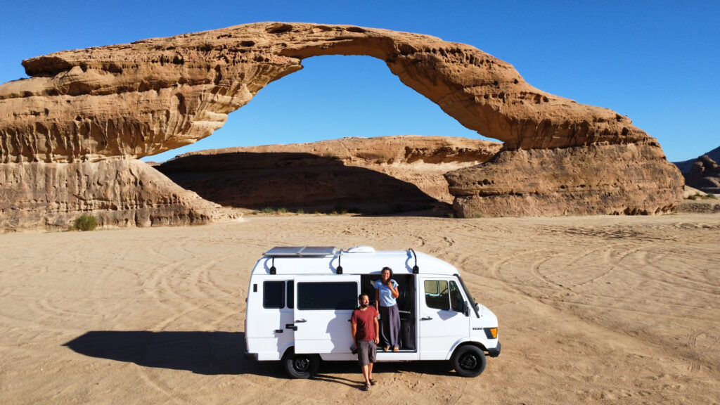 A man and woman standing beside a van in a vast desert landscape, with sand dunes and a clear blue sky in the background.