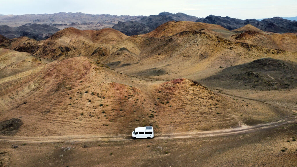 Aerial view of a white van driving through a vast, sandy desert landscape under a clear blue sky.
